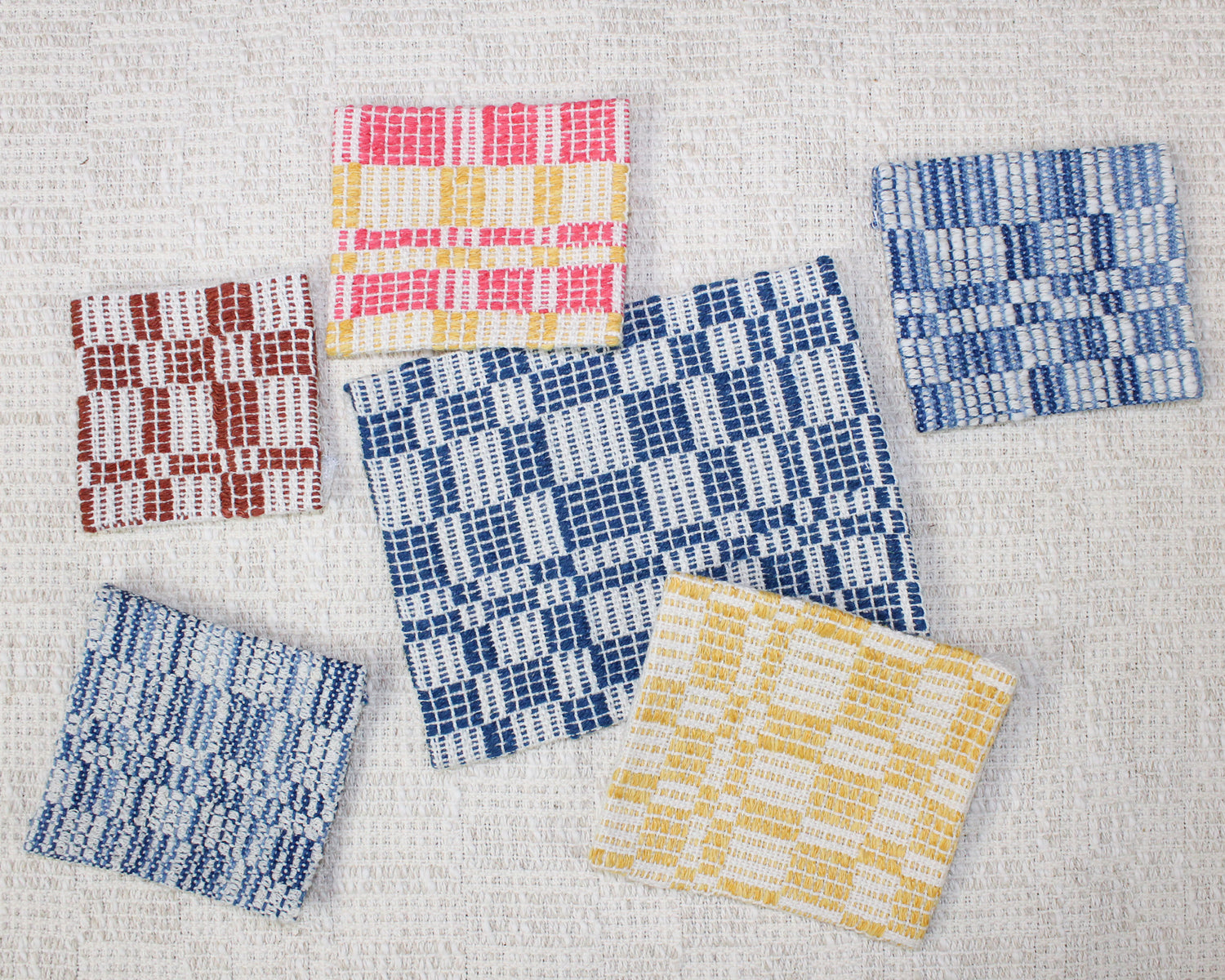several handwoven patches in shades of blue and yellow and pink sit on a neutral background. They all feature a grid woven design with lots of texture.