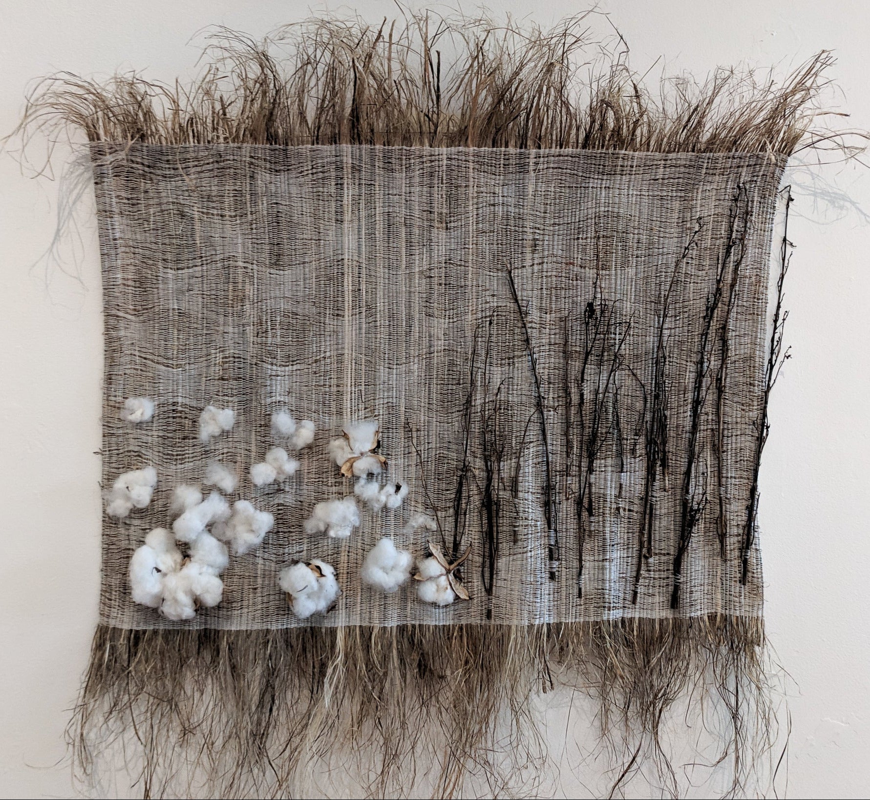 An image of a handwoven art work. It seems to be made of a rough grassy fiber, and features natural cotton bolls and hemp stems.