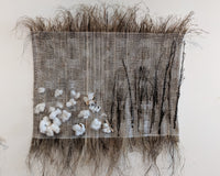 An image of a handwoven art work. It seems to be made of a rough grassy fiber, and features natural cotton bolls and hemp stems.