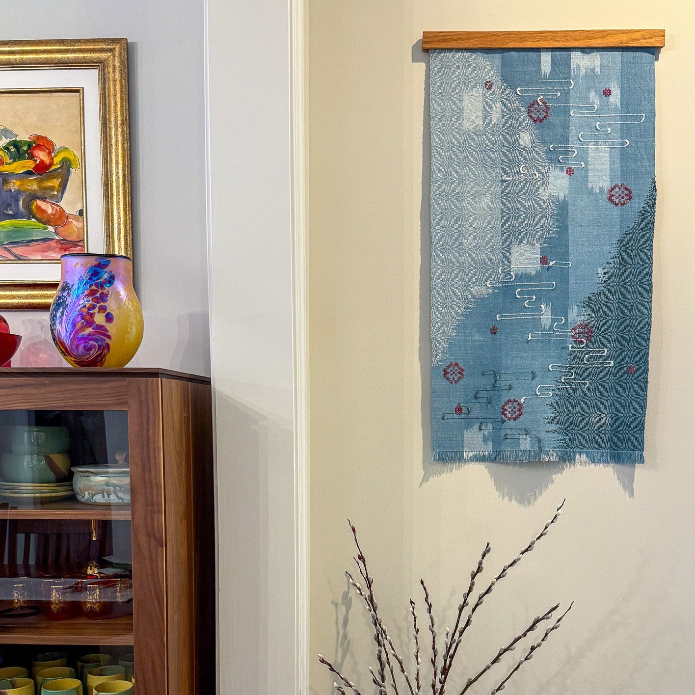 Decorative blue fabric with patterns hanging on a wall next to a cabinet with matching vase and dried flowers.