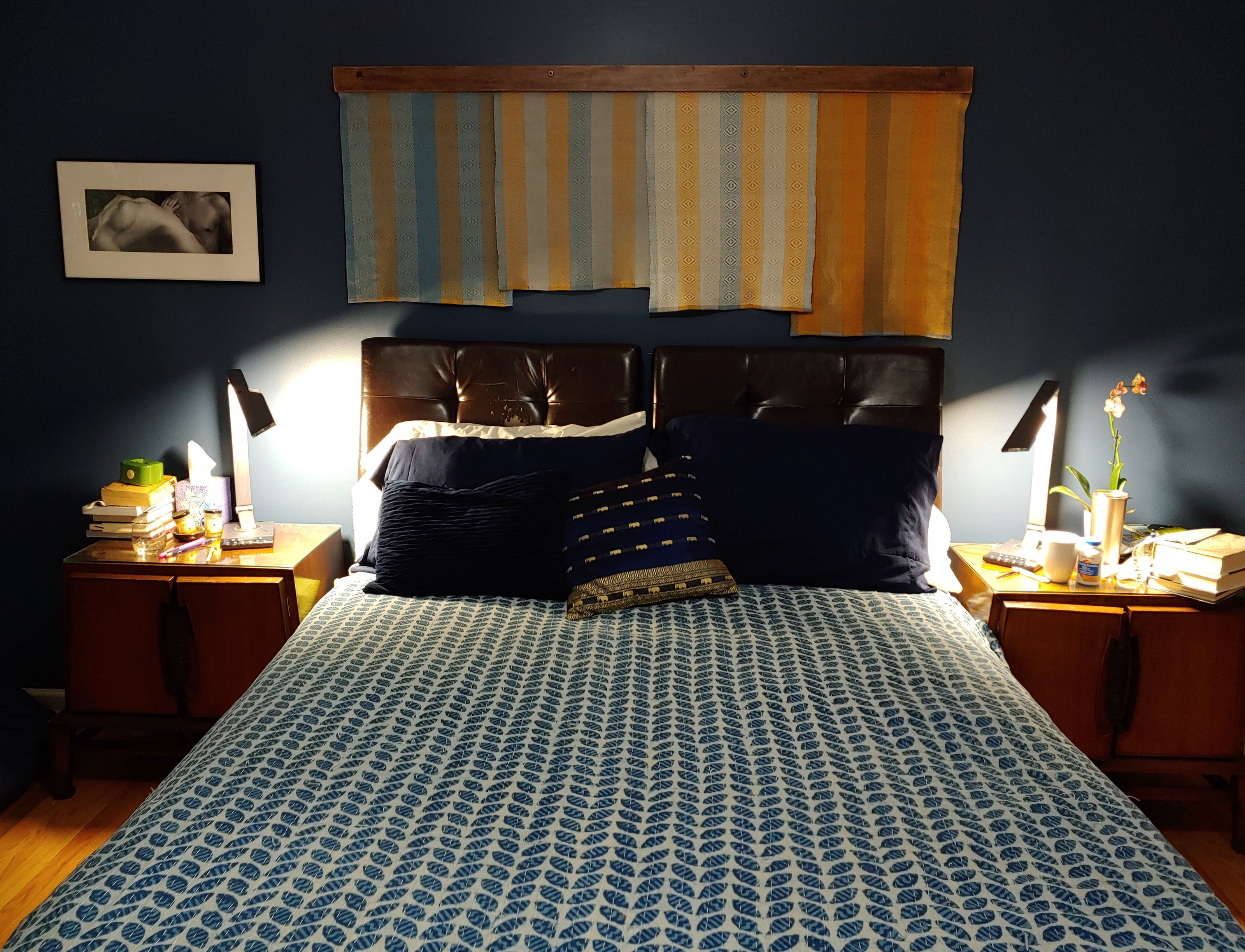 A bedroom scene with dark blue walls and patterned bedspread. On the wall behind the bed hangs a large textile hanging in stripes of blue and yellow. 